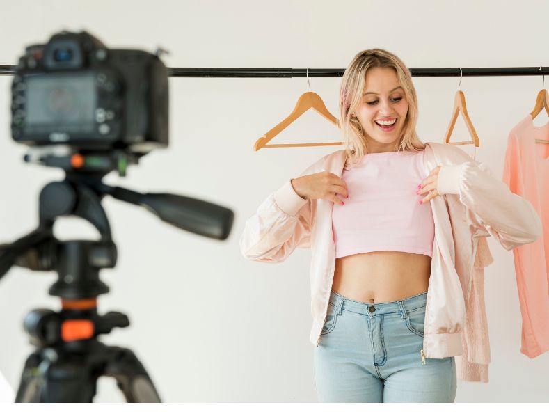 foto de uma influenciadora digital com uma camera a filamando enquanto mostra um look com blusa rosa e cala jeans